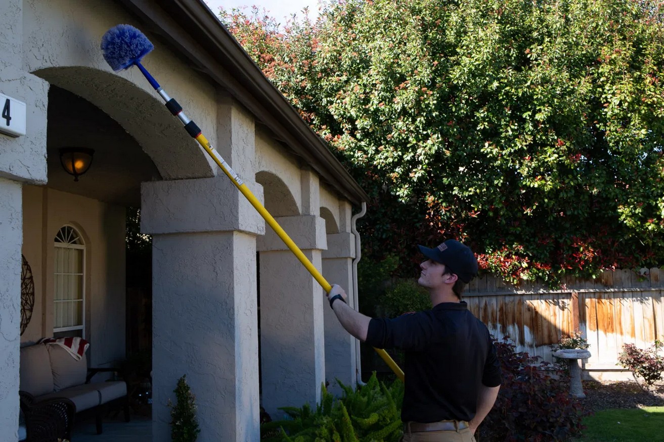 Pest control technician removing spider webs from home eaves