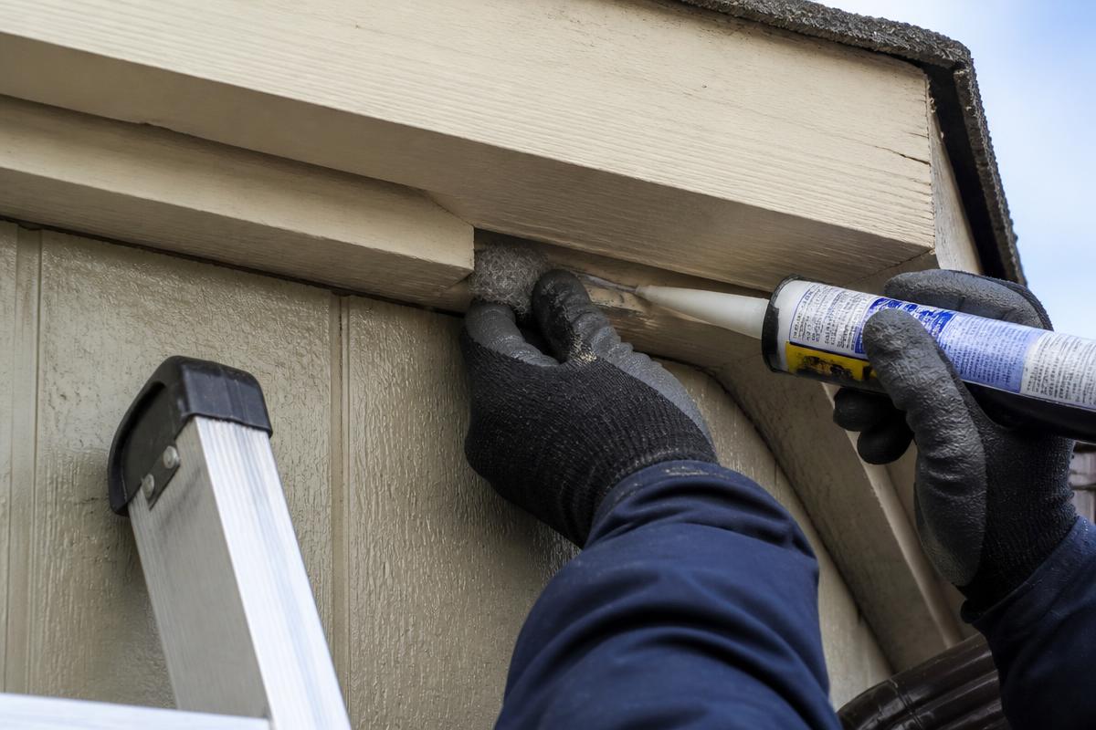 Gloved hands sealing a gap at a home's roofline with steel wool and caulk to block rodent entry